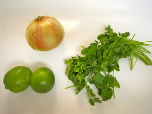 Whole yellow onion, fresh cilantro, and two limes arranged on a white surface as ingredients for carne asada tacos.