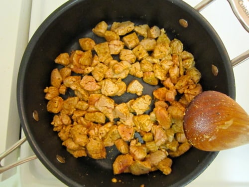 Cubed pork tenderloin browning in a skillet with a wooden spoon.