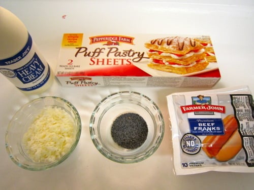 Ingredients laid out on a white surface including heavy cream, puff pastry sheets, shredded cheese, a bowl of poppy seeds, and a package of beef franks.