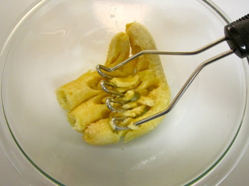 Mashed ripe bananas being pressed with a potato masher in a glass mixing bowl during bread preparation.
