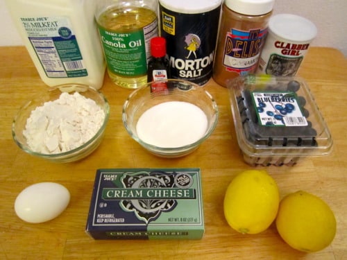 Ingredients for blueberry lemon cream cheese muffins arranged on a wooden surface, including milk, canola oil, salt, cinnamon, baking powder, vanilla extract, flour, sugar, blueberries, cream cheese, lemons, and an egg.