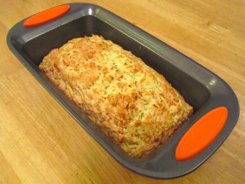 Baked zucchini parmesan loaf in a nonstick loaf pan with orange silicone handles, resting on a wooden surface.