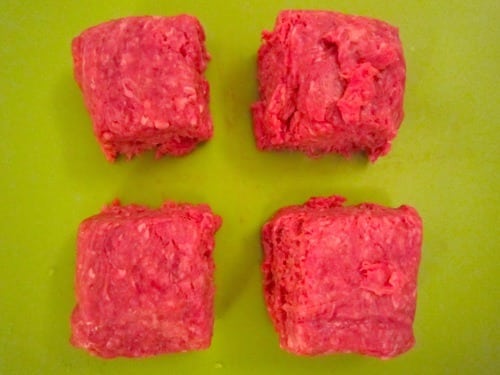 Four square-shaped portions of raw ground beef on a green cutting board, prepared for making burgers.