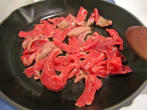 Thinly sliced beef cooking in a black skillet, prepared for beef and scallion stir-fry.