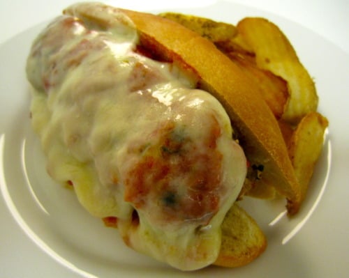 Meatball sub sandwich topped with melted cheese and marinara sauce, served with a side of potato chips on a white plate.