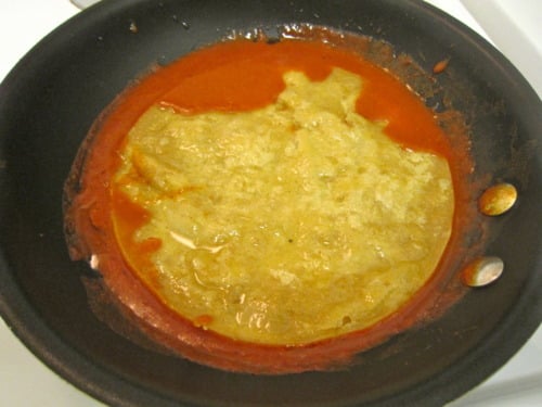 Corn tortilla coated in red enchilada sauce cooking in a skillet.