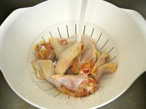Raw chicken wings rinsed under running water in a white colander before seasoning and baking.