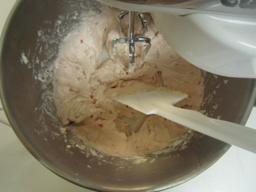 Mixing bowl with pale pink strawberry frosting being blended with a hand mixer and spatula inside.