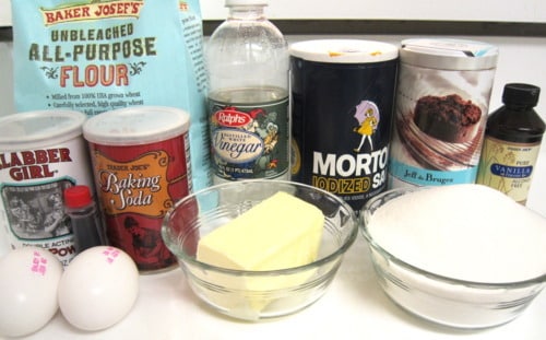 Ingredients for red velvet cupcakes, including flour, sugar, butter, eggs, cocoa powder, baking soda, baking powder, vinegar, vanilla extract, and salt, arranged on a counter.
