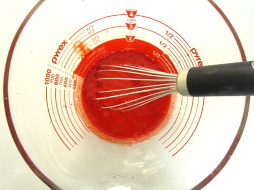 Red food coloring mixture being whisked in a glass measuring cup, prepared for red velvet cupcake batter.