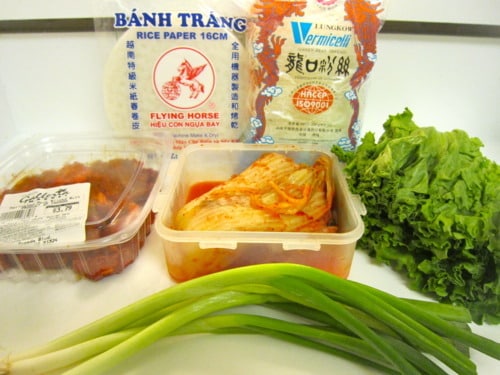 Ingredients for pork bulgogi and kimchi spring rolls arranged on a white surface, including rice paper sheets, dried vermicelli noodles, sliced marinated pork, kimchi, green onions, and fresh leafy greens.