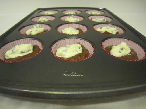 Muffin tin filled with double chocolate muffin batter topped with cheesecake and chocolate chips, ready for baking.