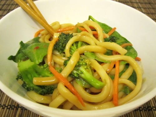 Bowl of udon noodles with broccoli, spinach, and carrot strips tossed in peanut sauce, with chopsticks resting on top