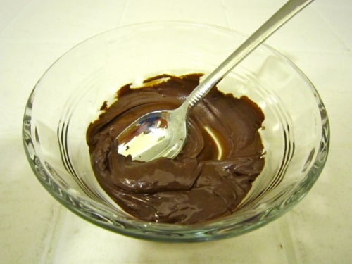 Smooth chocolate filling in a clear glass bowl with a spoon resting inside, prepared for a strawberries and cream birthday pie.