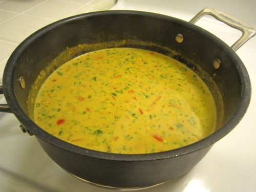 Thai coconut curry sauce simmering in a saucepan with visible herbs and red bell peppers, prepared for broiled tilapia.