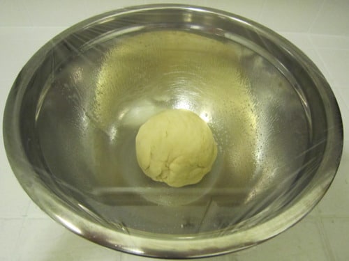 Pizza dough formed into a smooth ball resting in a lightly oiled metal bowl and covered with plastic wrap, ready to rise.