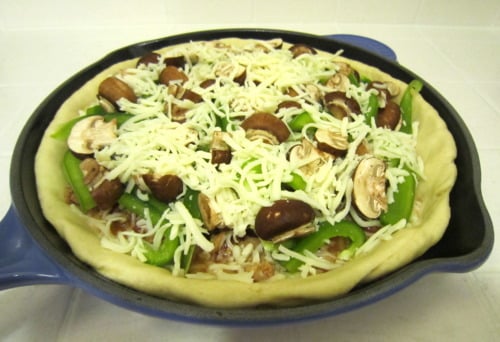 Unbaked deep dish sausage pizza topped with mushrooms, green peppers, and shredded cheese in a cast iron skillet.