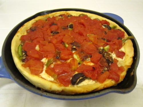 A skillet deep-dish pizza topped with chunky tomato pieces, melted cheese, vegetables, and sausage, baked inside a cast-iron pan.