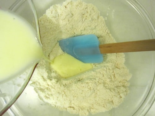 Milk mixture being poured into a bowl of dry ingredients with a blue spatula for making Irish soda bread muffins.
