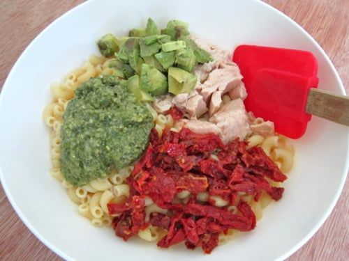 Macaroni pasta in a white bowl with pesto, diced avocado, tuna, and sun-dried tomatoes, ready to be mixed.