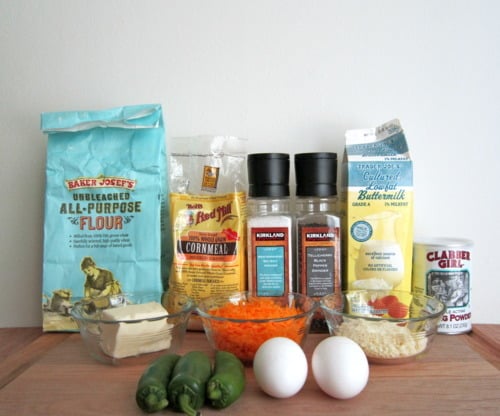 Ingredients for jalapeño cheddar parmesan cornbread arranged on a table, including flour, cornmeal, baking powder, buttermilk, spices, shredded cheese, jalapeños, eggs, and butter.