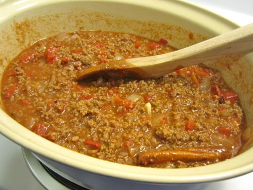 Bolognese sauce with ground meat, diced tomatoes, onions, garlic, and whole cinnamon sticks simmering in a pot with a wooden spoon.