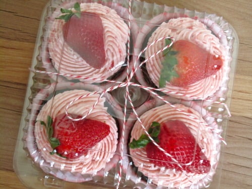 Chocolate cupcakes topped with pink strawberry frosting and fresh strawberries, packaged in a clear container with twine.
