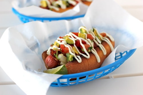 Bacon-wrapped Sonoran hot dog topped with diced tomatoes, onions, avocado, and a drizzle of mayonnaise, served in a blue basket lined with parchment paper.