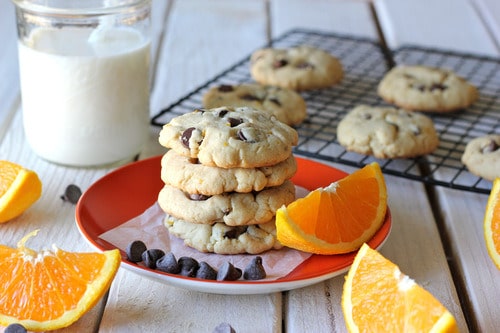 Stack of orange chocolate chip cookies on a red plate with orange slices, chocolate chips, and a glass of milk, with more cookies cooling on a rack in the background.