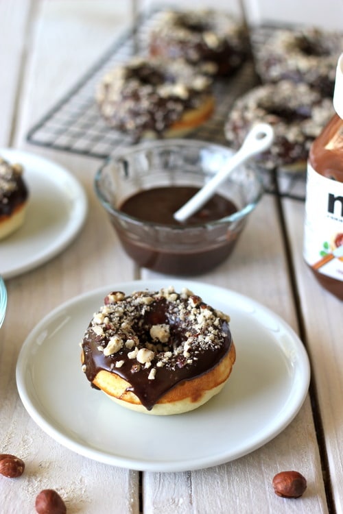 Nutella-glazed donuts topped with chopped hazelnuts on a plate, with more donuts and chocolate glaze in the background.