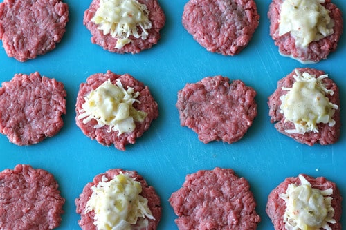 Raw ground beef patties arranged on a blue cutting board, some topped with green chili con queso filling, prepared for stuffed cheeseburger sliders.