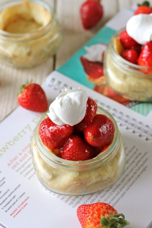Individual strawberry pie in a glass jar topped with fresh strawberries and a dollop of whipped cream, surrounded by whole strawberries and an open cookbook.