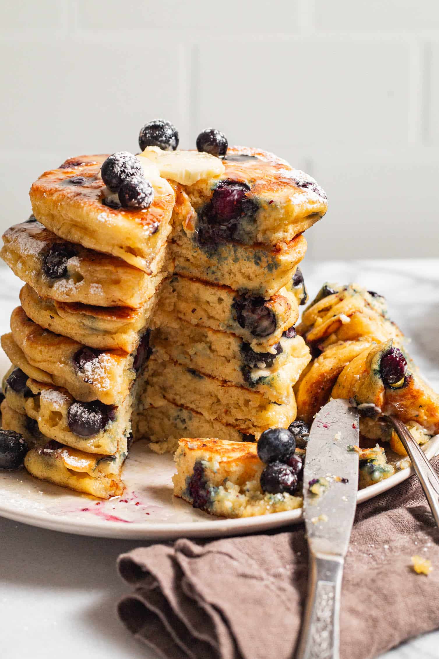 Stack of fluffy blueberry pancakes topped with butter and fresh blueberries, served on a white plate with a knife and fork.