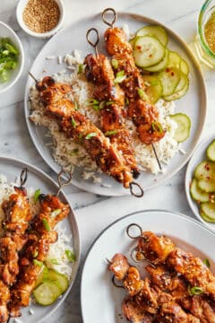 Plates of marinaded, sesame chicken kabobs, served on a bed of white rice with marinated cucumbers.