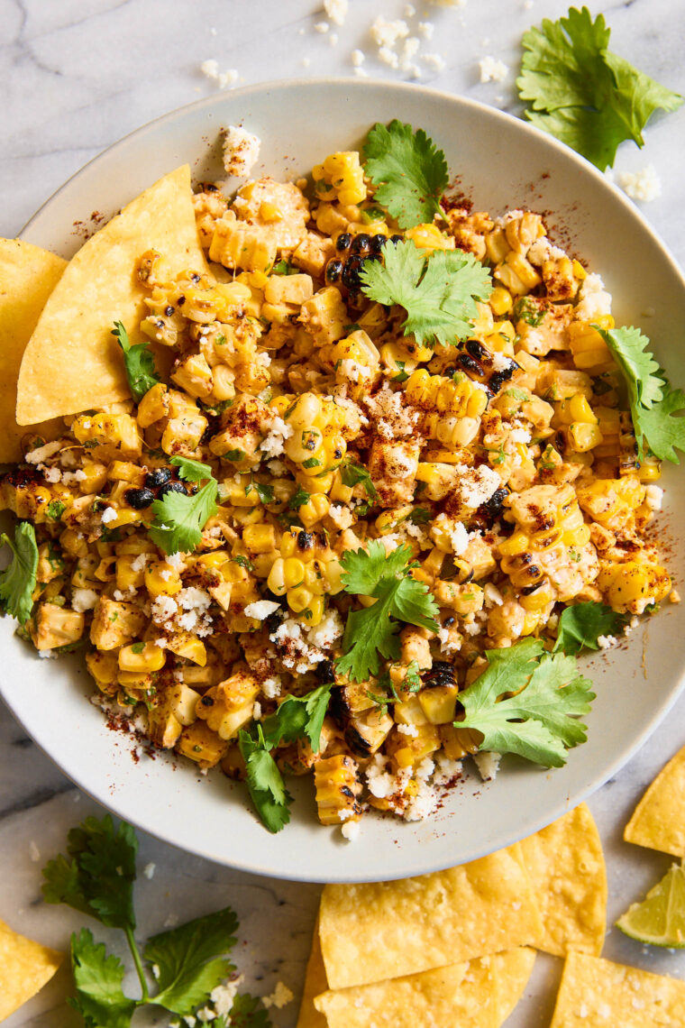 Tortilla chip scooping creamy Mexican corn dip with roasted corn, herbs, crumbled cheese, and chili powder, with bowl and chips in background.